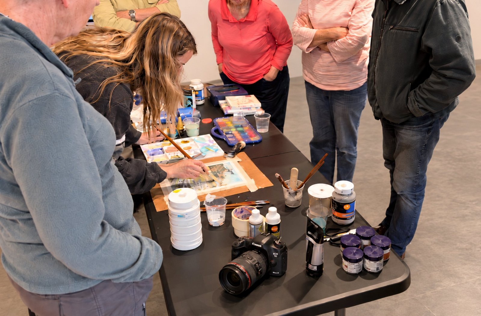 Samantha Lee Osner painting at the studio table, a camera beside her, guests watching